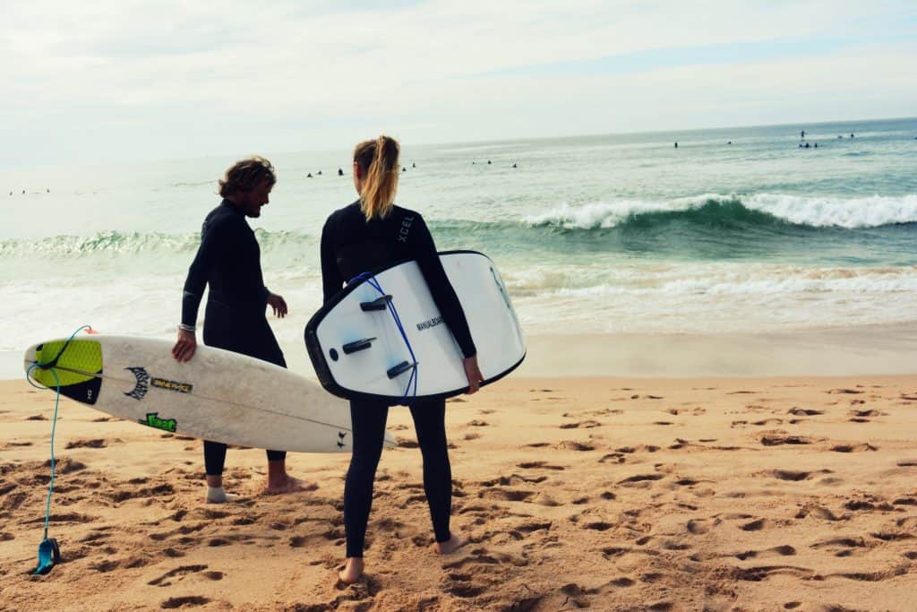 Two individuals wearing surfing wetsuits on a sandy beach.